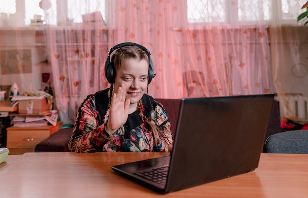 Una niña discapacitada con ojos rasgados se sienta en una mesa con auriculares y se comunica mientras mira una computadora portátil. formación de personas con discapacidad