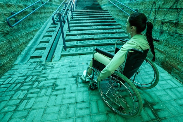 Una niña discapacitada sentada en una silla de ruedas en el fondo de las escaleras. Los días grises de los discapacitados, problemas de movimiento, enfermedades.