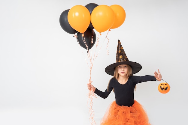 Una niña con un disfraz de bruja de carnaval y un sombrero sosteniendo una canasta para golosinas y sustos de globos naranjas y negros