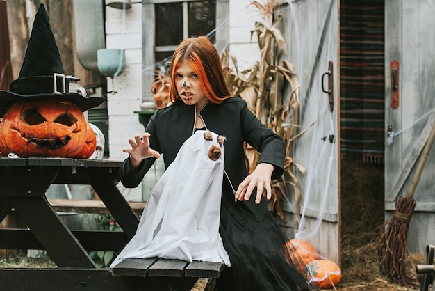 Foto una niña con un disfraz de bruja con un perro disfrazado de fantasma divirtiéndose en el porche de una casa decorada para celebrar una fiesta de halloween