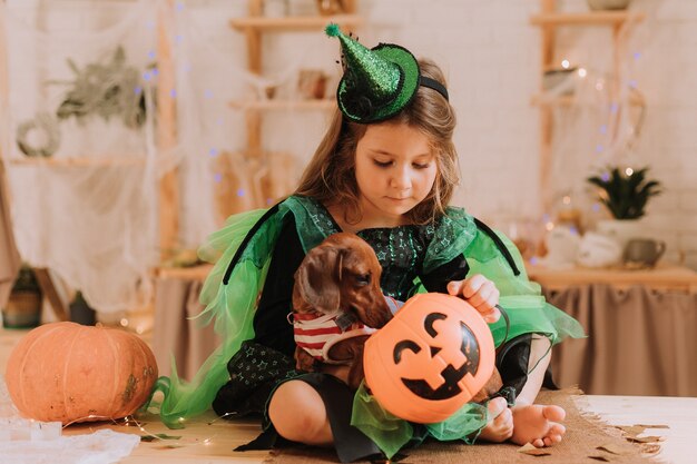 Niña en un disfraz de Carnaval de Halloween y un perro salchicha están sentados en la cocina