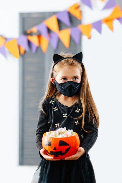 Una niña con un disfraz de Halloween con una máscara está sosteniendo una calabaza llena de golosinas