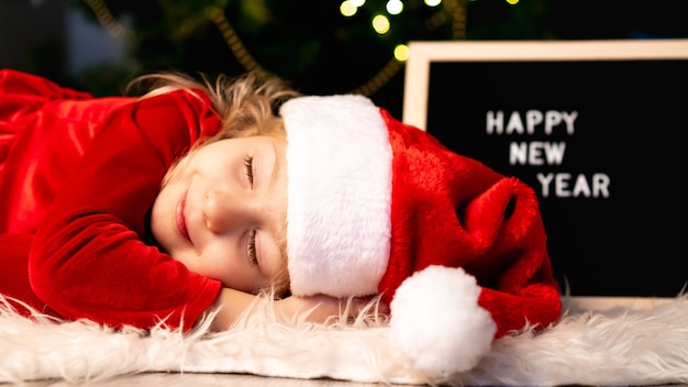 Una niña con un disfraz de Navidad y un gorro de Papá Noel duerme bajo el árbol esperando un milagro.