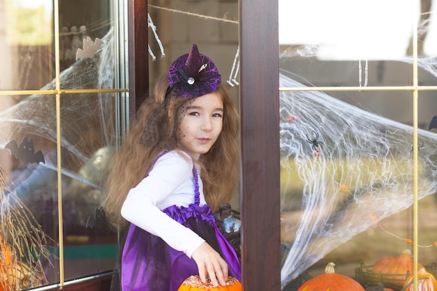 Una niña disfrazada de bruja en el alféizar de la ventana está jugando con una telaraña y una araña decorando la casa para las vacaciones de Halloween Terrible paisaje miedo y calabazas de terror