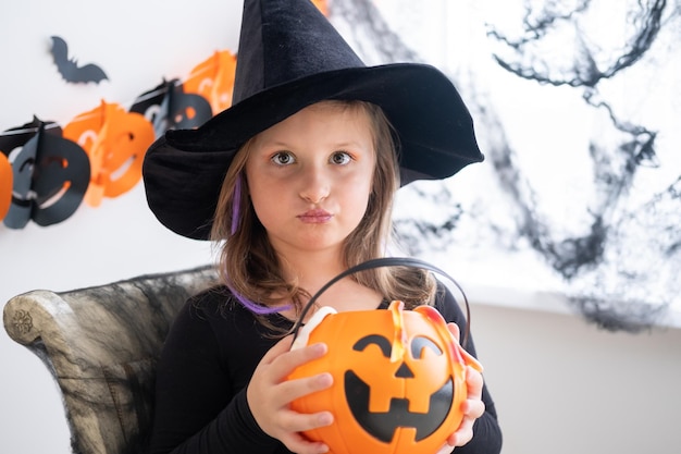 niña disfrazada de bruja sosteniendo gato de calabaza con dulces, celebrando Halloween en casa