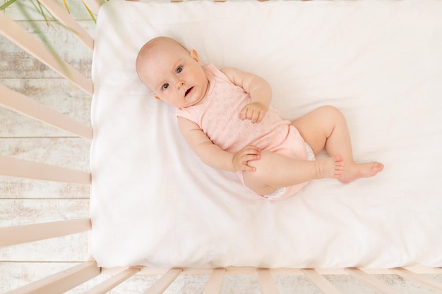 Niña divertida en una cuna con un traje rosa de seis meses en una cama de algodón blanco se despierta por la mañana o se va a la cama