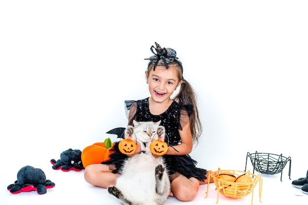 Niña divertida en un disfraz de bruja para Halloween con un gato en un traje en sus manos