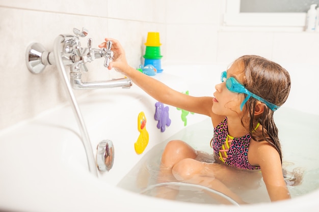 Niña divertida en gafas de baño azules mira a la cámara y sonríe encantadoramente mientras vierte el agua de la ducha sobre ella