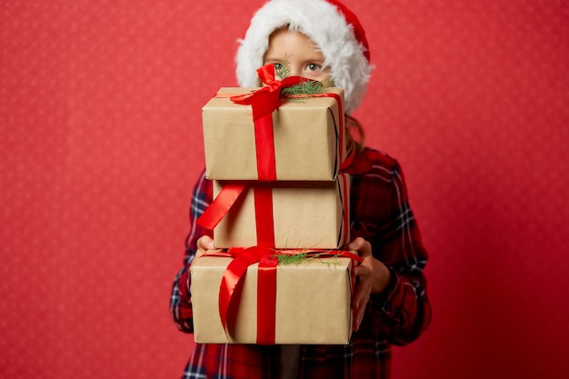 Niña divertida con gorro de Papá Noel con muchas cajas de regalo de Navidad aisladas sobre fondo rojo,