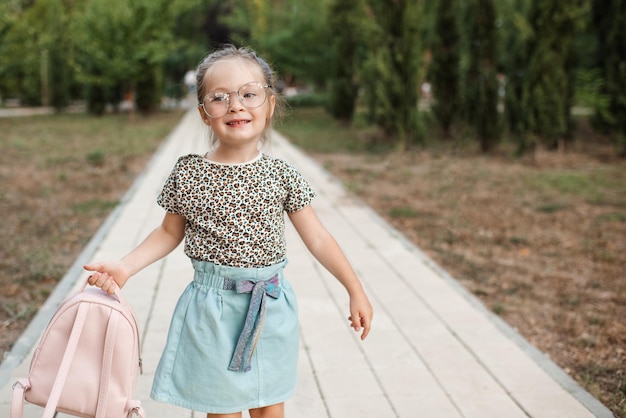 Una niña divertida y linda de 4 a 5 años usa anteojos y ropa informal con mochila en el parque