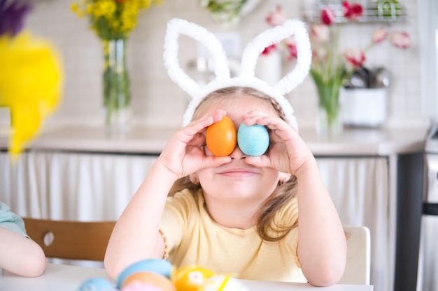 Niña divertida con orejas de conejo en la cabeza cubriendo los ojos con huevos de Pascua multicolores en casa