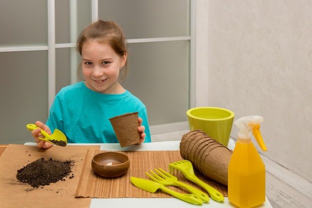 Niña divertida preparando la tierra para plantar semillas herramientas de plantación cerca de macetas de turba aspersor con agua