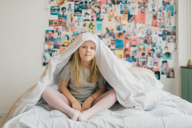 Niña divertida que se sienta en la cama blanca y que sostiene la manta sobre su cabeza. Retrato del estilo de vida del niño femenino rubio sonriente feliz que se cubre con la manta en fondo decorativo de la pared.