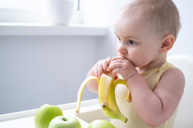 niña divertida sentada en una silla para niños comiendo plátano en la cocina blanca