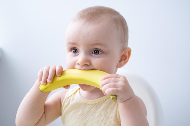 niña divertida sentada en una silla para niños comiendo plátano en la cocina blanca