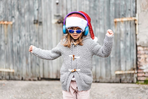 Niña divertida con sombrero de papá noel y gafas de sol bailando contra un fondo grunge de madera al aire libre en navidad feliz navidad año nuevo concepto de infancia en los días de navidad