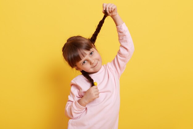 Niña divertida tirando de coletas y mirando directamente a la cámara con una linda sonrisa, vestida con camisa rosa pálido, niño encantador contra la pared amarilla.