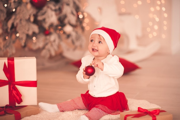 Niña divertida con traje de santa claus jugando con decoración navideña en la habitación con luces. Temporada de invierno.