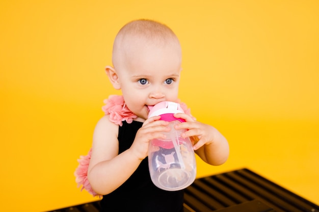 Niña divertida vestida con un traje de baño negro y rosa, sombrero grande sentado en la tumbona de madera con una botella de agua sobre fondo amarillo. Concepto de vacaciones de verano.