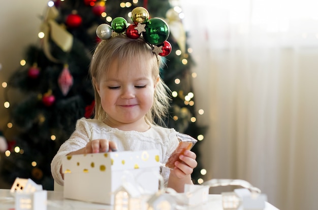 Niña divertida en vestido blanco abriendo caja de regalo de Navidad en casa cerca del árbol de Navidad