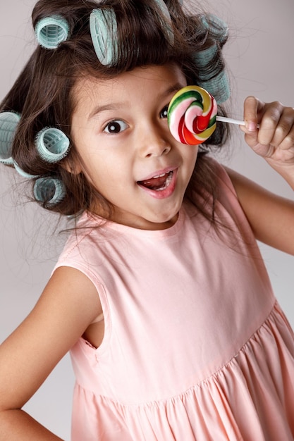 Niña divertida en vestido rosa y rizadores de pelo con piruleta sobre fondo gris.