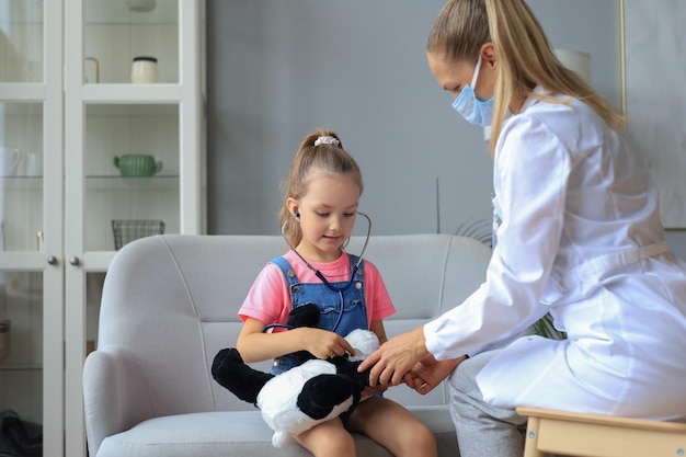 Niña en el doctor para un chequeo Doctor juguetonamente revisando el latido del corazón de un oso de peluche