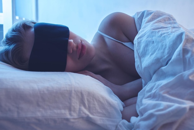 Niña dormida con una máscara para dormir sobre una almohada ortopédica con iluminación nocturna, ropa de cama blanca.