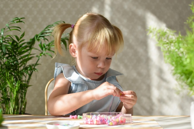 Una niña de dos años está sentada recogiendo cuentas de pequeñas cuentas Desarrollo de habilidades motoras finas en un niño Bricolaje