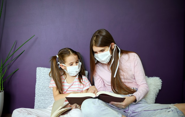 Una niña de dos en máscara médica emplazamiento en el piso de madera y leyendo libros durante la cuarentena