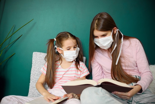 Una niña de dos en máscara médica emplazamiento en el piso de madera y leyendo libros durante la cuarentena