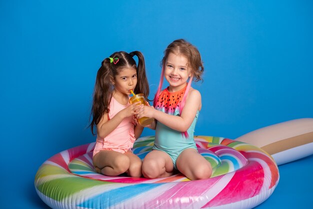 Niña de dos niños felices en traje de baño bebiendo jugo de naranja sentado en un colchón inflable de colores.