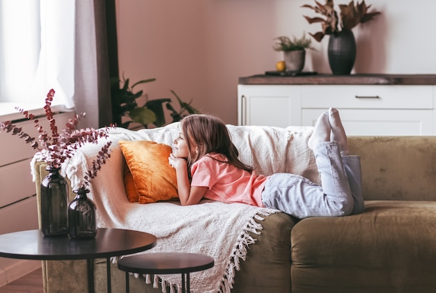 Una niña en edad escolar se acuesta en un sofá en una habitación luminosa y mira por la ventana. Los sueños de los niños