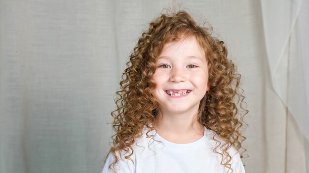 Niña en edad preescolar de pelo castaño rojizo rizado en camiseta blanca se ve recta y sonríe con el diente perdido posando para la cámara en primer plano de fondo gris