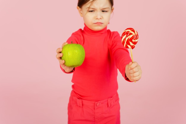 La niña elige entre una piruleta y una manzana verde. El concepto de nutrición adecuada. Un niño en una pared rosa tiene un dulce de azúcar en la mano y una manzana. Dificultad de elección