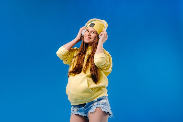 Una niña embarazada con una chaqueta amarilla y auriculares se encuentra sobre un fondo azul.