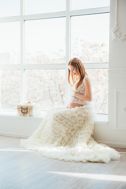 Niña embarazada pelirroja con un vestido blanco cerca de la ventana