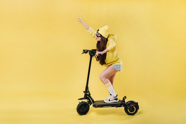 Una niña embarazada en ropa amarilla en un scooter eléctrico sobre un fondo amarillo aislado.