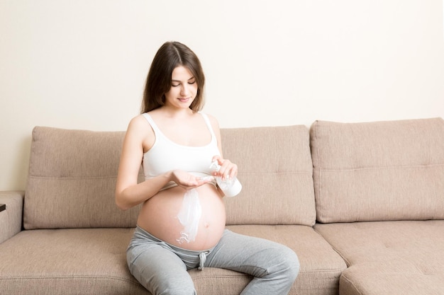 Una niña embarazada se sienta en casa en la cama y frotis sonríe con una crema antiestrías en el estómago Concepto de preparación y expectativa de la maternidad durante el embarazo