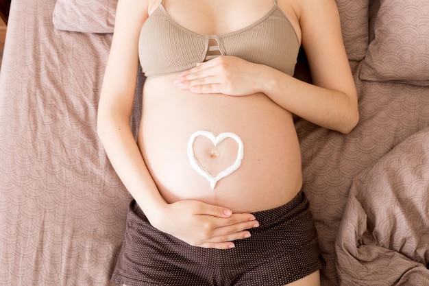 Una niña embarazada se sienta en su casa en la cama y se mancha el corazón con una crema antiestrías en el estómago Concepto de preparación y expectativa de la maternidad durante el embarazo