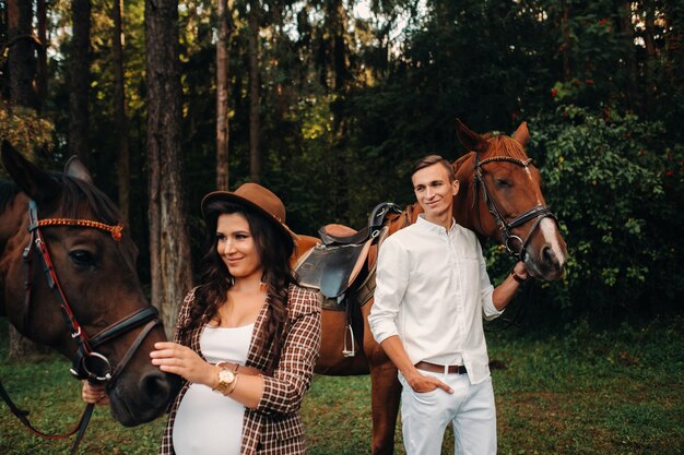 Una niña embarazada con un sombrero y un hombre vestido de blanco junto a los caballos en el bosque en la naturaleza