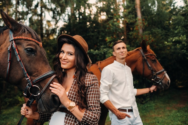 Una niña embarazada con un sombrero y un hombre vestido de blanco de pie junto a los caballos en el bosque en la naturaleza. Elegante mujer embarazada con su marido con caballos. Pareja casada.