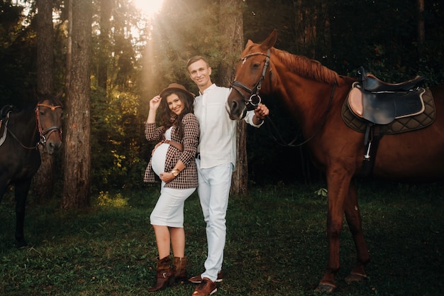 Una niña embarazada con un sombrero y un hombre vestido de blanco de pie junto a los caballos en el bosque en la naturaleza. Elegante mujer embarazada con su marido con caballos. Pareja casada.