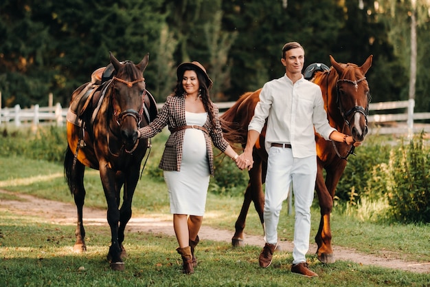 Una niña embarazada con un sombrero y su esposo con ropa blanca se encuentran junto a los caballos en el bosque en la naturaleza.