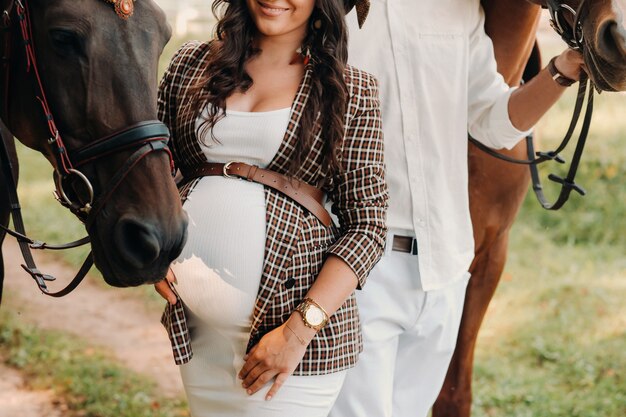 Foto una niña embarazada con un sombrero y su esposo con ropa blanca están junto a los caballos en el bosque en la naturaleza