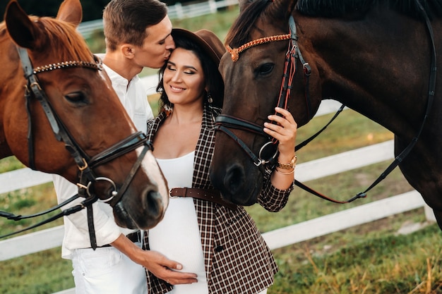 Una niña embarazada con un sombrero y su esposo vestido de blanco se paran junto a los caballos cerca del corral de caballos. Elegante mujer embarazada con un hombre con caballos. Familia.