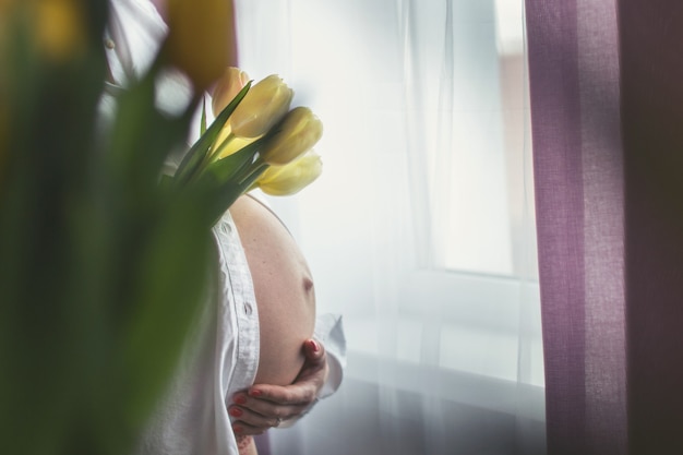 Niña embarazada sosteniendo flores mujer embarazada de pie junto a la ventana con flores