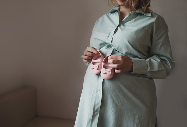 Una niña embarazada sostiene pantuflas para niños pequeños en una pared gris. esperando el nacimiento de un niño