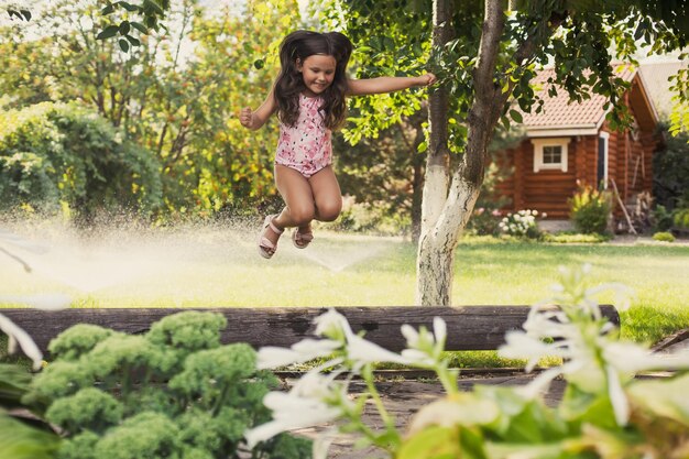 Niña emocionada jugando afuera saltando de un tronco mirando hacia abajo en el patio trasero complaciendo con árboles y rociadores de agua en el fondo durante el día Soñando con convertirse en gimnasta deportiva