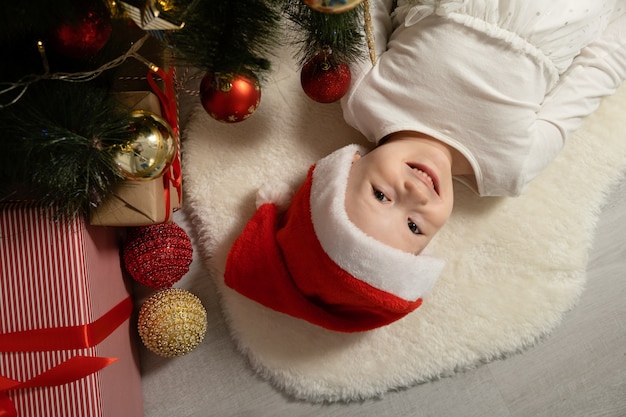 niña emocionada con sombrero de Santa tumbado bajo el árbol de Navidad. vista superior.