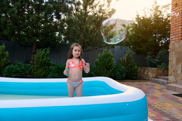 Una niña encantadora atrapa pompas de jabón que sopla en una piscina de agua inflable en el jardín de su casa en un día soleado de verano